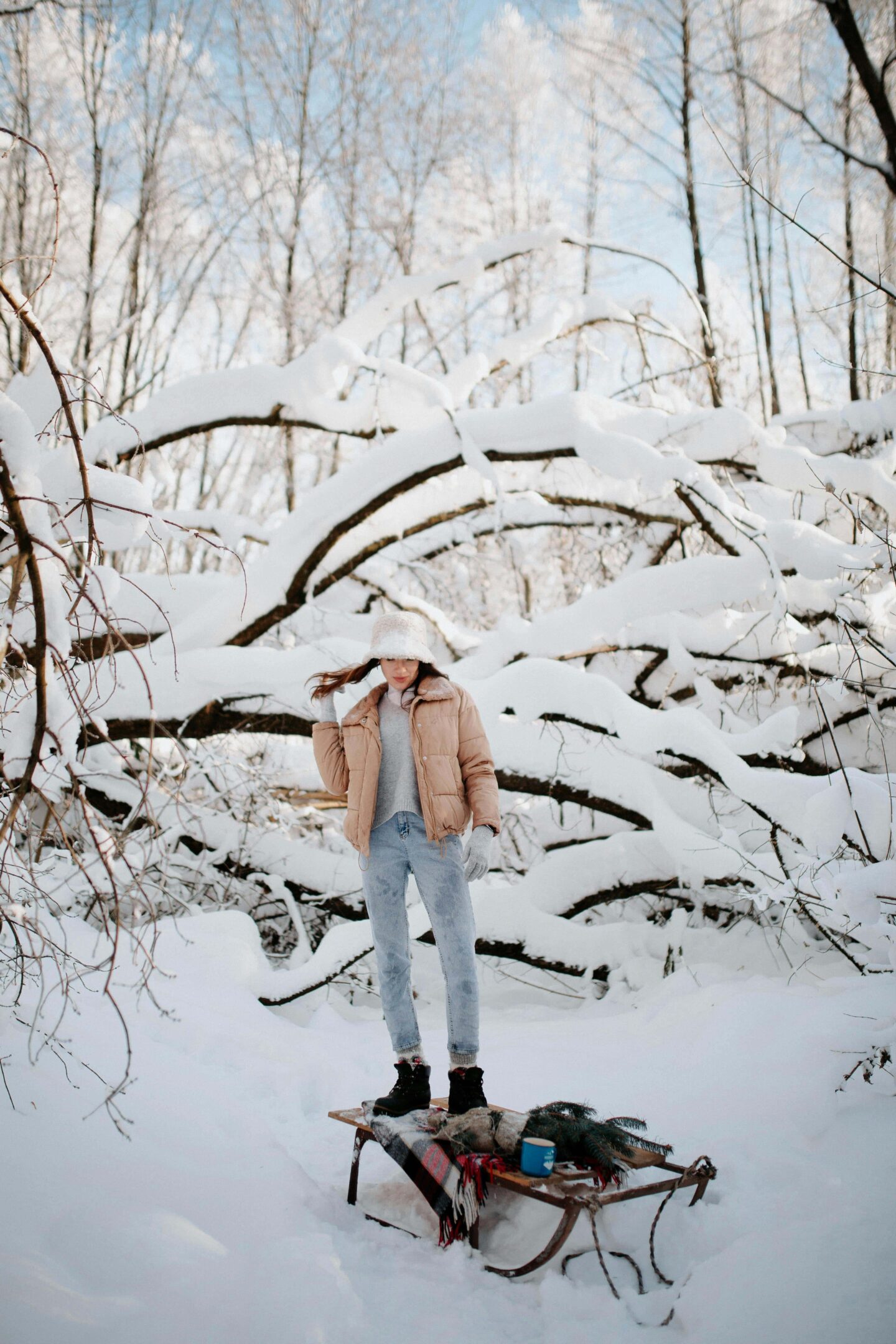 71 Best Apres Ski Party Outfit Ideas for 2025 – The Ultimate Guide! A woman in a stylish apres ski outfit poses in a snowy forest, wearing a beige puffer jacket, light-wash jeans, black winter boots, and a faux fur hat—perfect for a cozy yet chic apres ski party look.